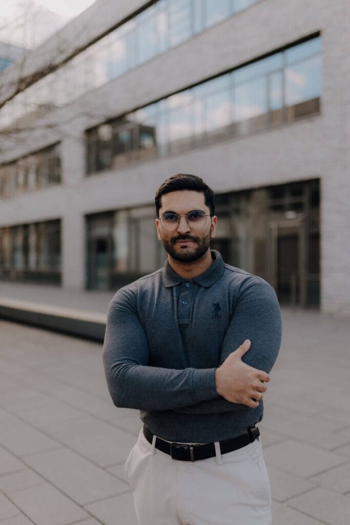 Ein Mann mit Brille und Bart steht im Freien vor einem modernen Gebäude. Er trägt ein dunkles, langärmeliges Polohemd und eine helle Hose und hat einen Arm vor der Brust verschränkt.