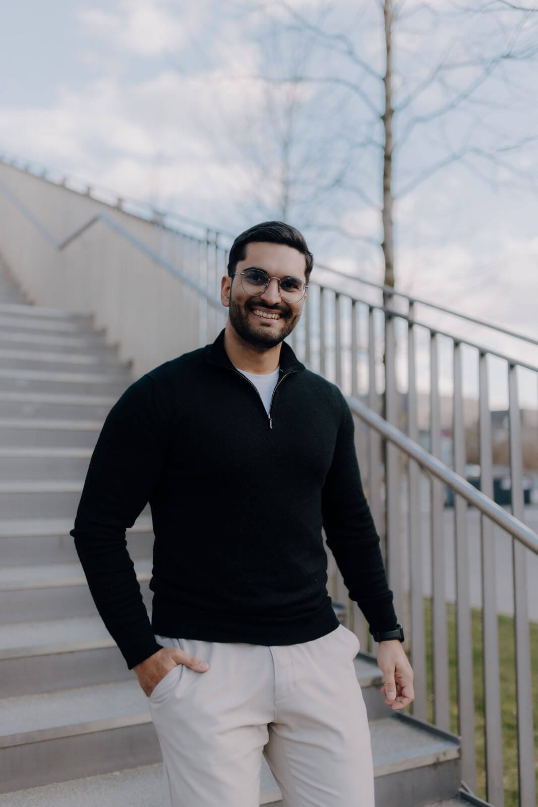 Ein Mann, der eine Brille, einen schwarzen Pullover und eine helle Hose trägt, steht draußen auf einer Treppe und lächelt, die Hände in den Taschen.