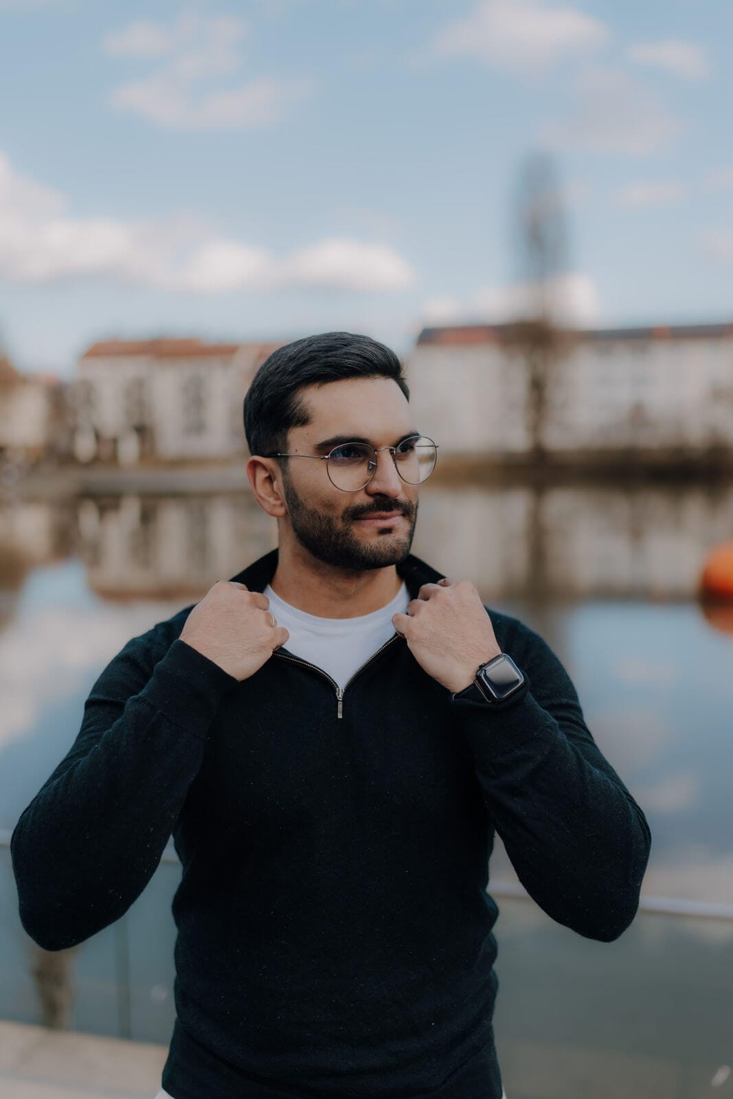 Ein Mann mit Brille und schwarzem Pullover steht im Freien an einem Gewässer und hält seinen Kragen mit beiden Händen fest. Im Hintergrund sind Gebäude und ein bewölkter Himmel zu sehen.
